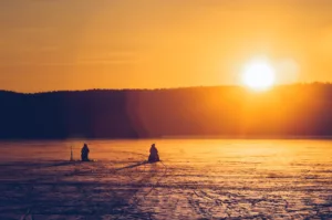 silhouette of two persons sitting while snow fishing on an iced covered body of water at dawn