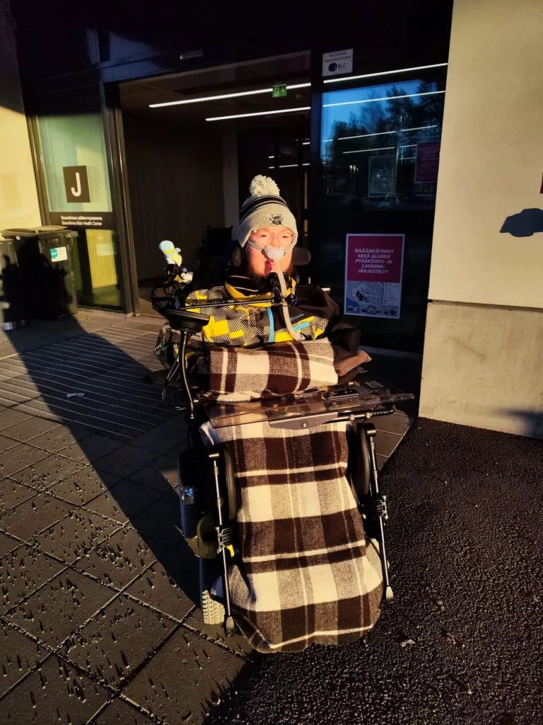 Savonlinnan sosiaali- ja terveyskeskuksen ovella viltteihin kääriytyneenä. Aurinko paistaa kirkkaalta taivaalta.