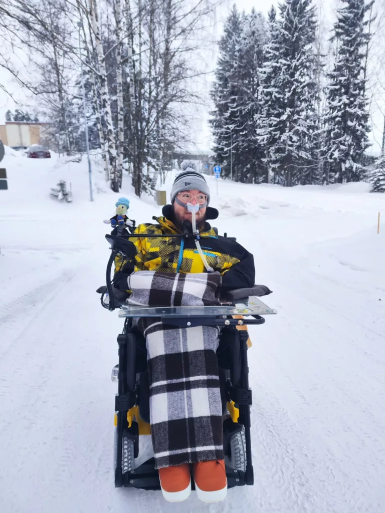 Minä pyörätuolilla meidän kotipihassa. Maisema on hyvin luminen.