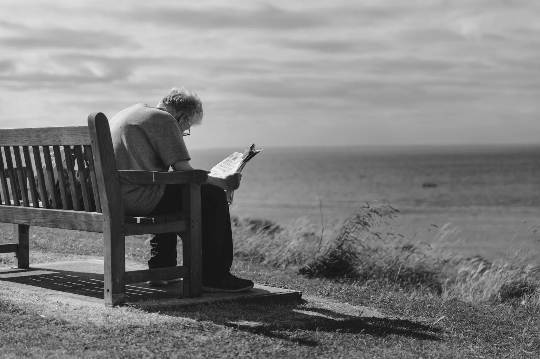 grayscale photo of man sitting on brown wooden bench reading news paper during day time